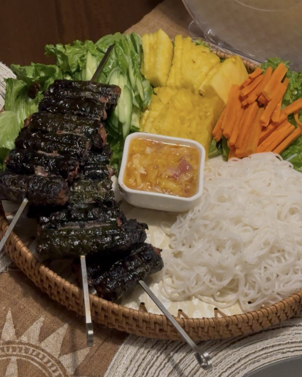 Grilled betel leaf rolls on skewers on a woven tray with lettuce, pineapple, carrots, rice noodles and dipping sauce
