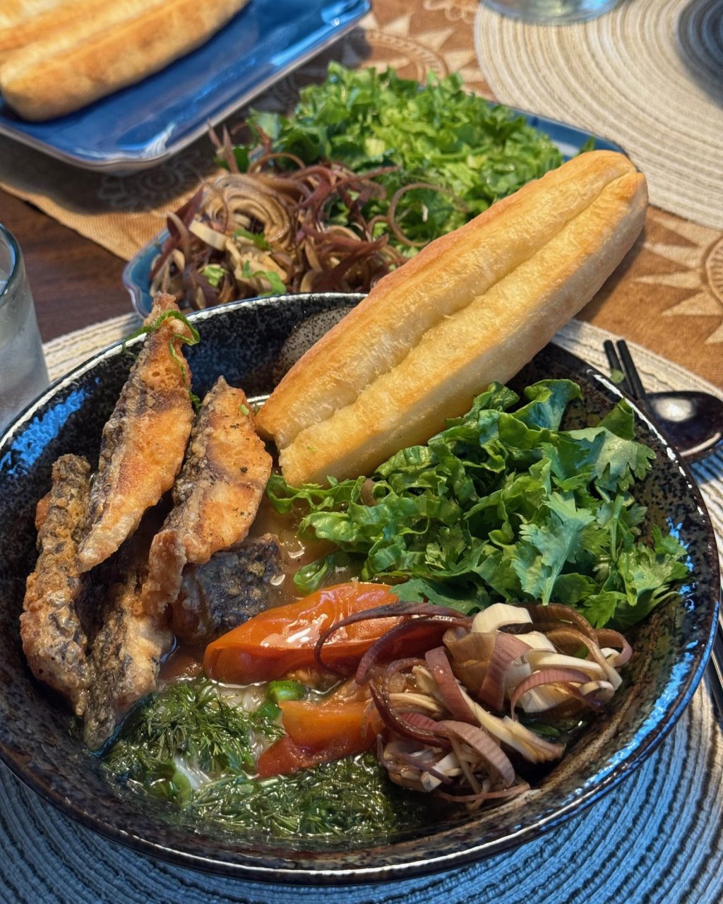 Bowl of fish noodle soup with crispy fried fish, tomatoes, shallots, fresh herbs and a Chinese doughnut