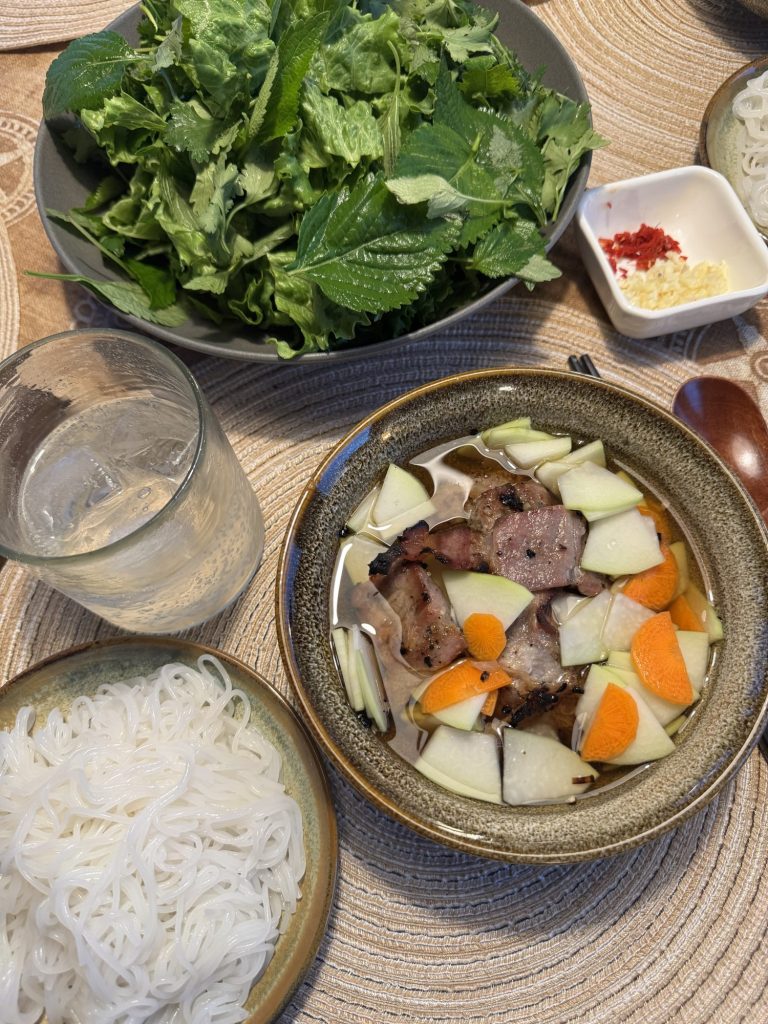 Bún chả served at the table: a bowl of grilled pork in dipping sauce with pickled papaya and carrot, a plate of rice vermicelli and a bowl of lettuce and herbs