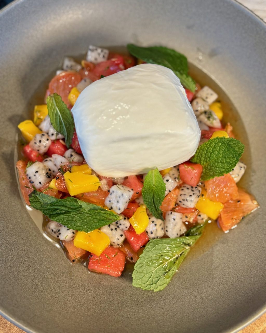 Overhead view of a whole burrata on diced watermelon, dragon fruit and mango with mint leaves in a gray bowl