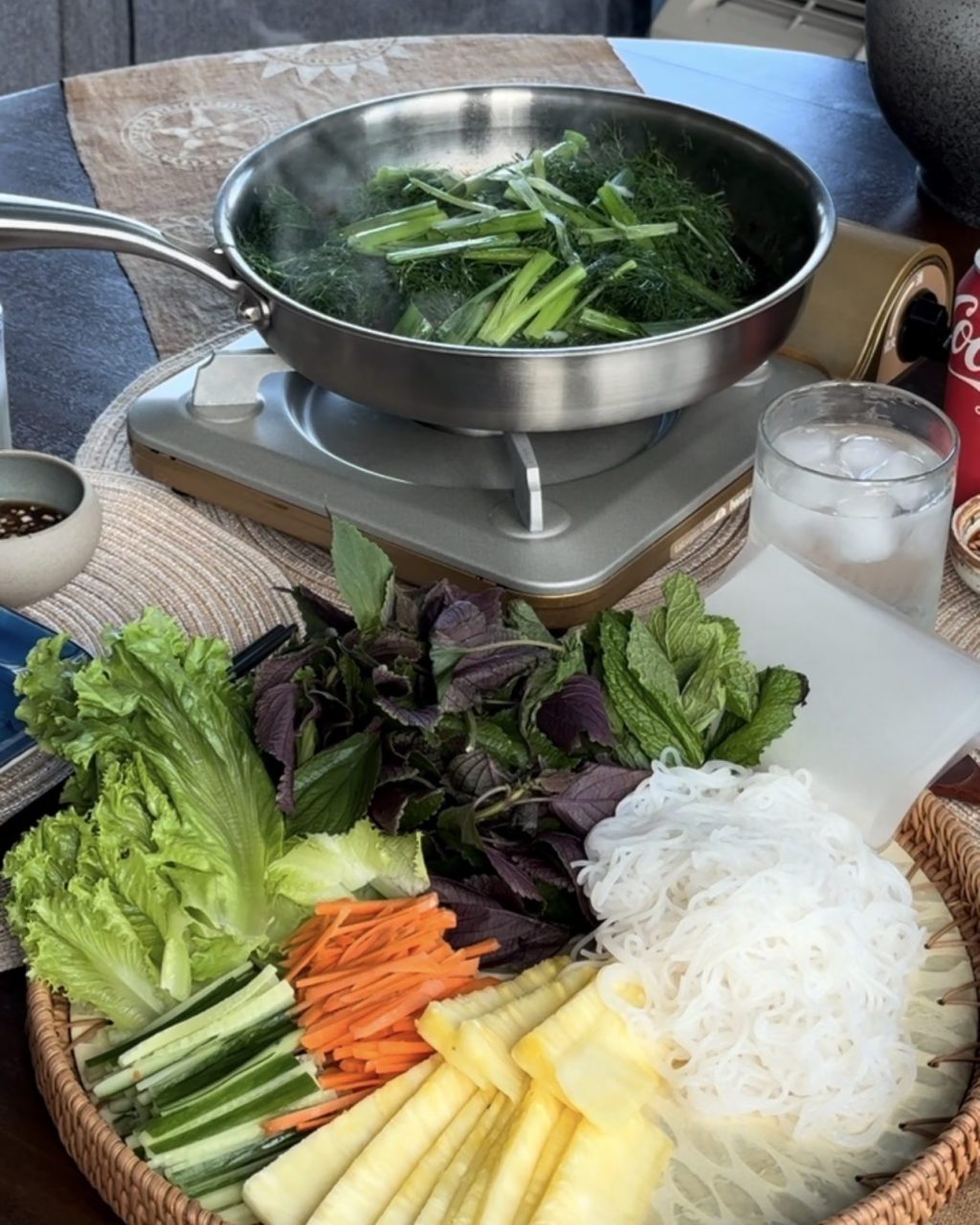 Pan of steamed fish topped with dill and green onions on a burner, beside a platter of lettuce, herbs, carrots, cucumber, pineapple and vermicelli