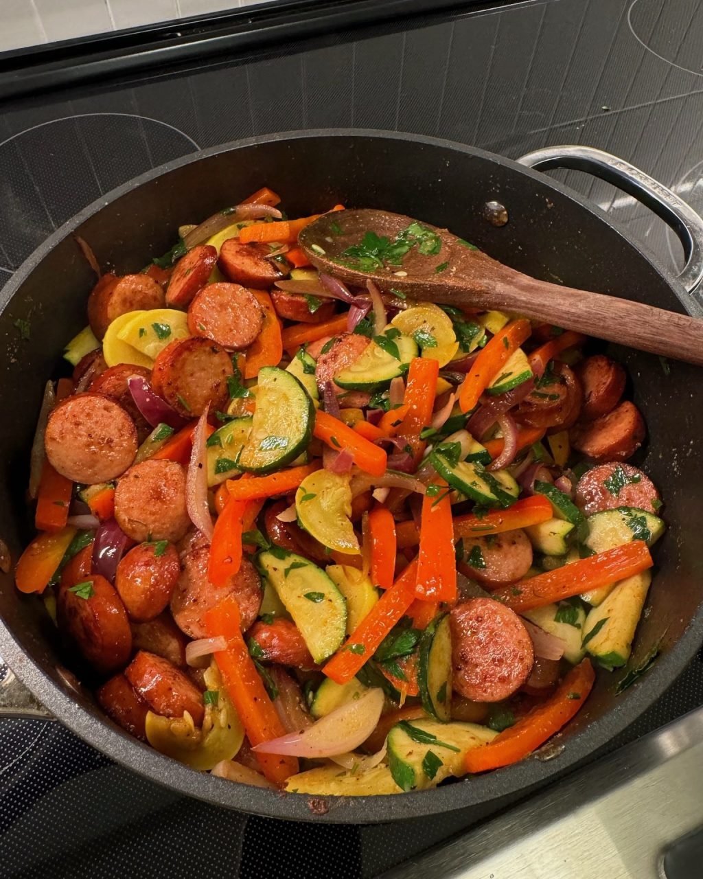 Cajun andouille sausage coins with zucchini, yellow squash, bell pepper strips and red onion in a large skillet, topped with parsley, wooden spoon resting on the side