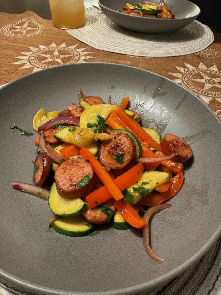 A plated serving of Cajun sausage and vegetables in a gray bowl, with a second bowl and a drink on the table behind