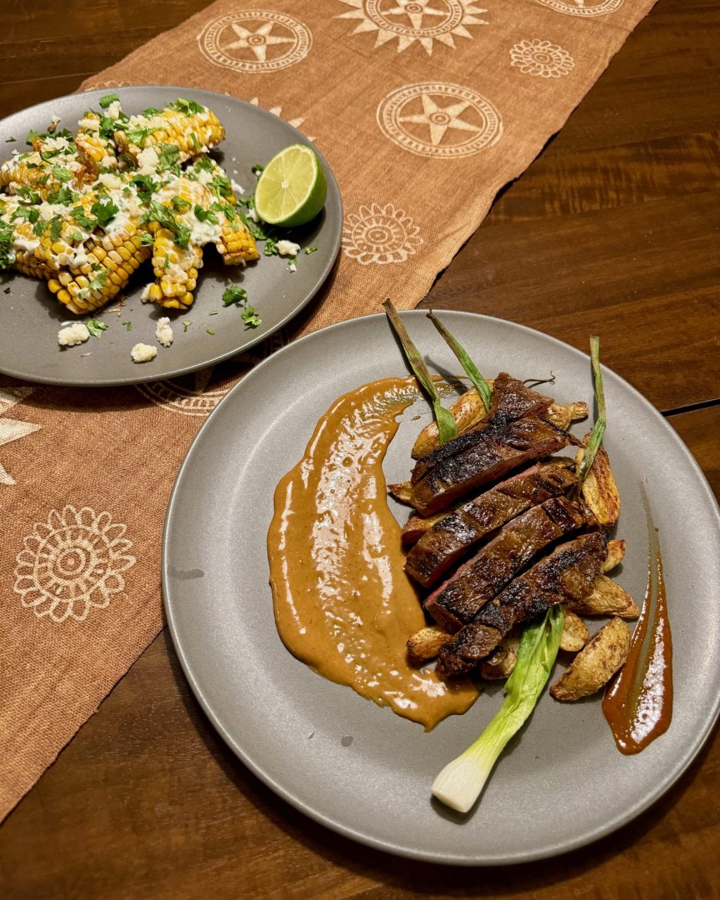 Sliced grilled carne asada with chile salsa, grilled scallions and potatoes on a grey plate, with a plate of street corn ribs behind