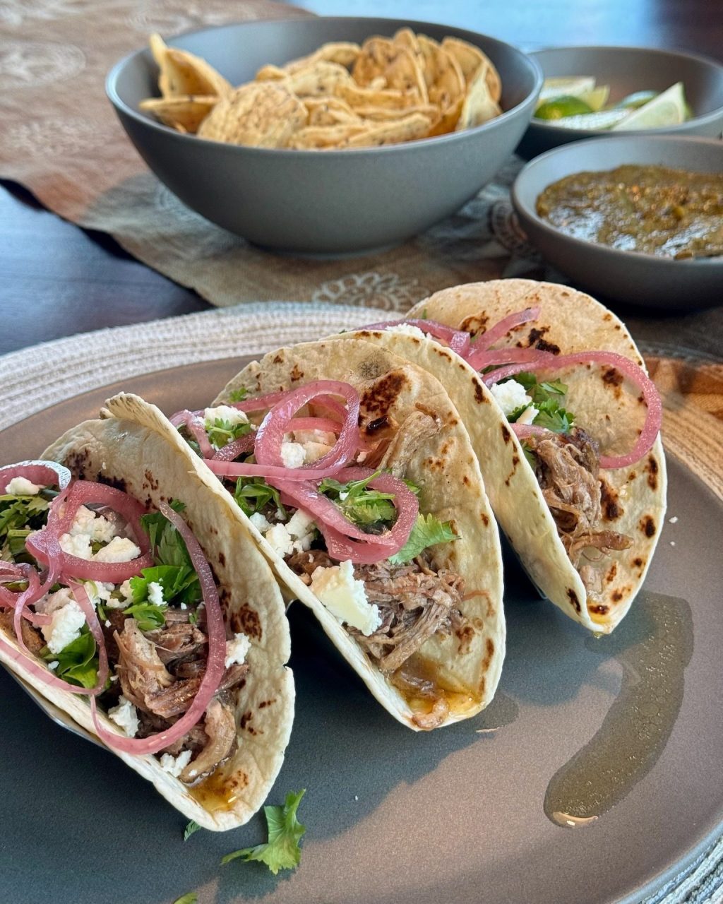 Three carnitas tacos topped with pickled red onion, crumbled cheese and cilantro on a grey plate, with chips and salsa behind