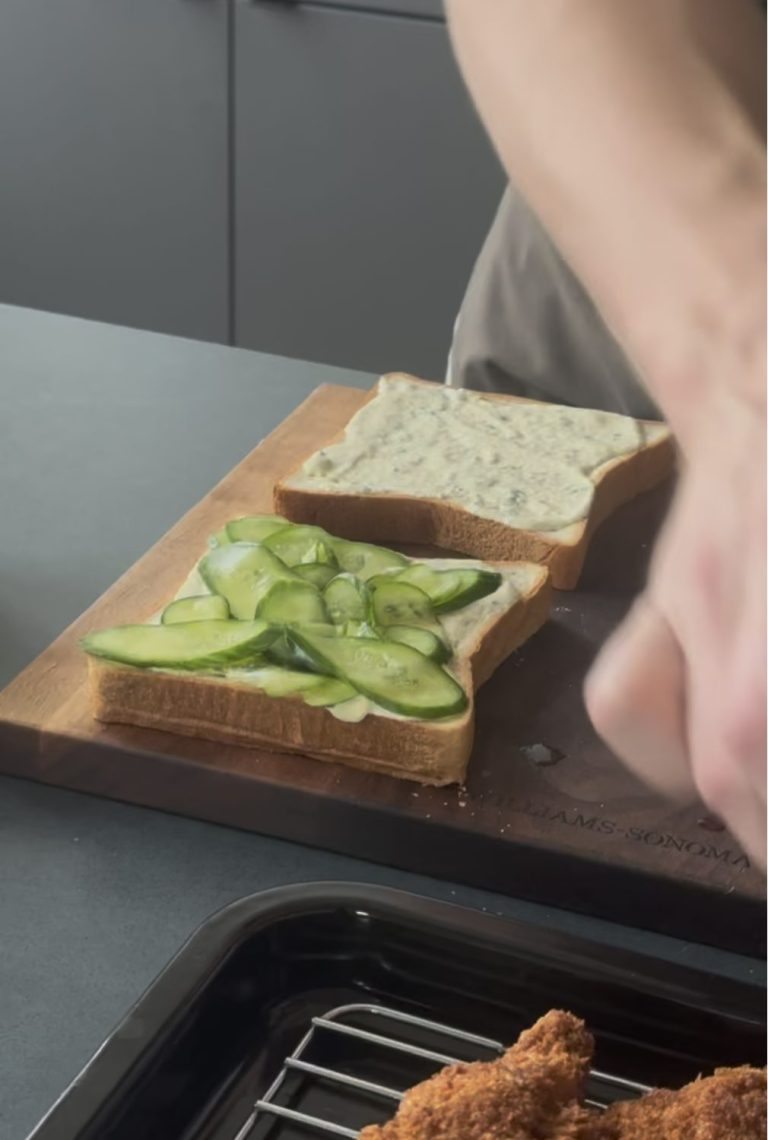 Two slices of white bread spread with jalapeño tartar sauce on a cutting board, one topped with thin salted cucumber slices
