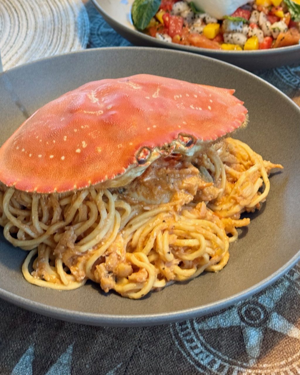 Crab tomato cream spaghetti in a gray bowl, topped with a whole red crab shell, with a burrata fruit salad behind