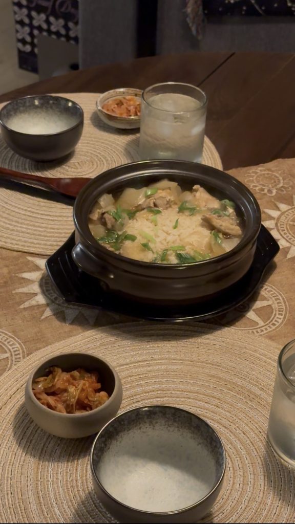 Korean chicken soup in a black clay pot with chicken, radish, rice and sliced green onions, with kimchi on the side