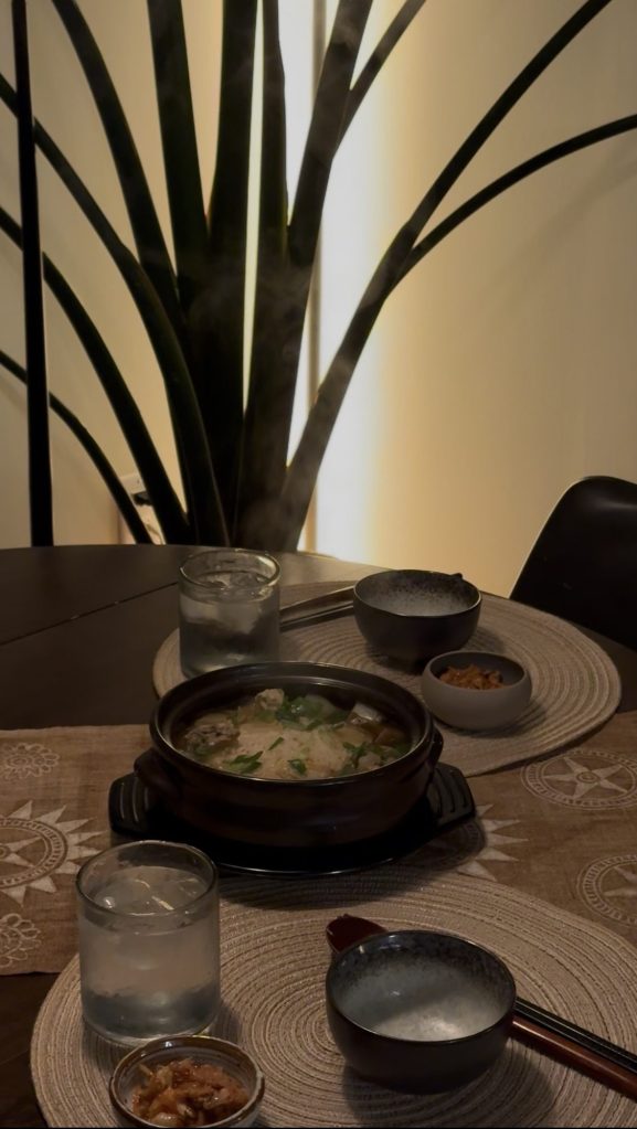 Steaming clay pot of Korean chicken soup on a dark table set for two, with rice bowls, kimchi and a tall plant behind in warm evening light