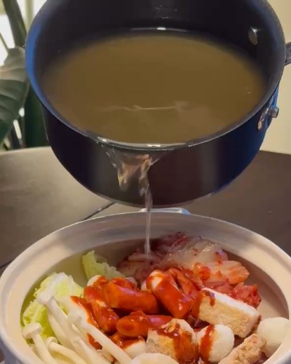 Golden dashi being poured from a saucepan into a clay pot filled with kimchi, rice cakes, fish cakes and mushrooms