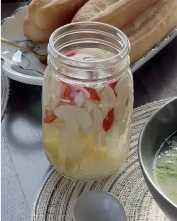 A mason jar of sliced garlic and red chilies in rice vinegar on the table next to a bowl of phở and fried dough sticks