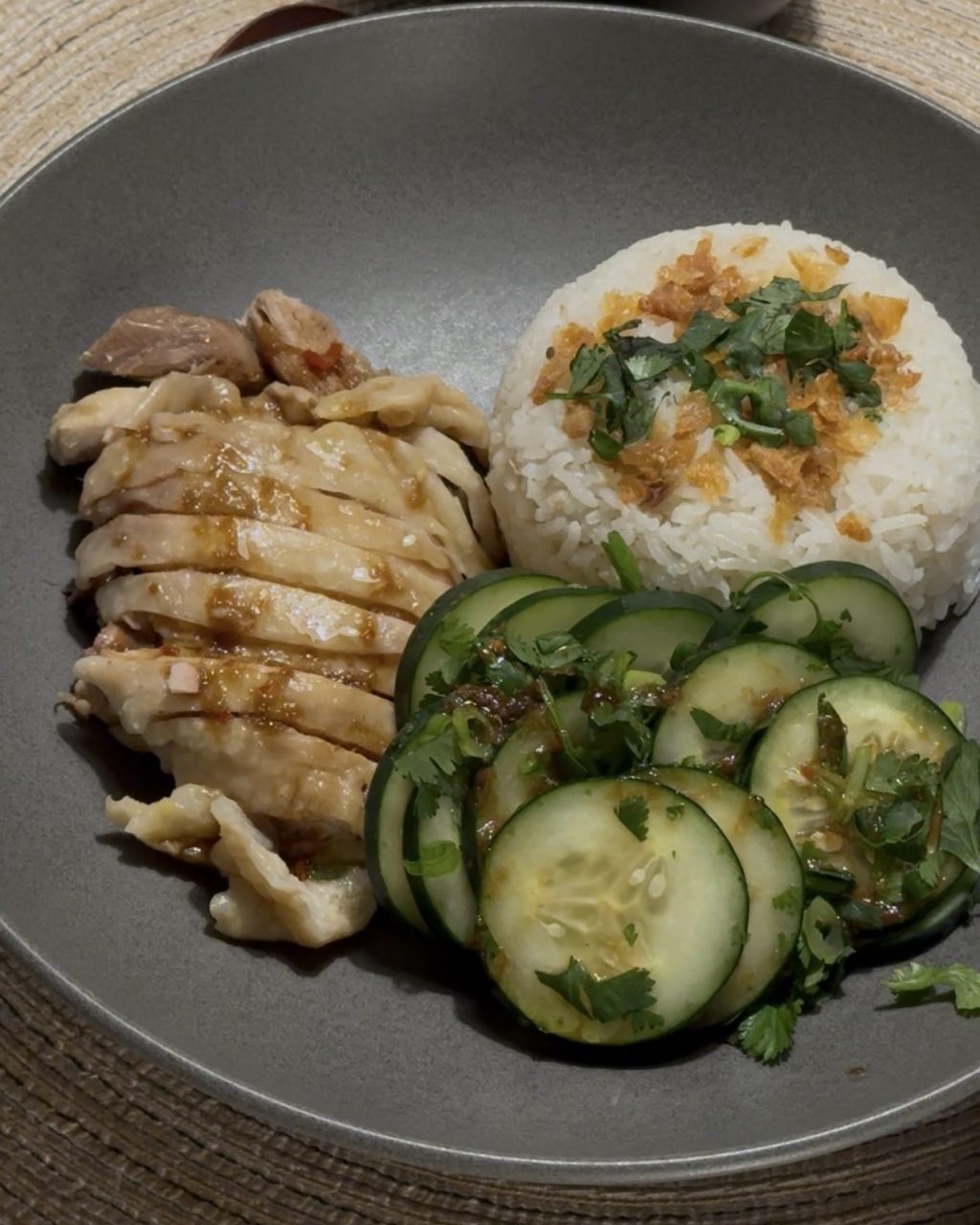 Sliced poached chicken with a mound of rice topped with crispy shallots, sliced cucumbers and cilantro, with a small bowl of broth