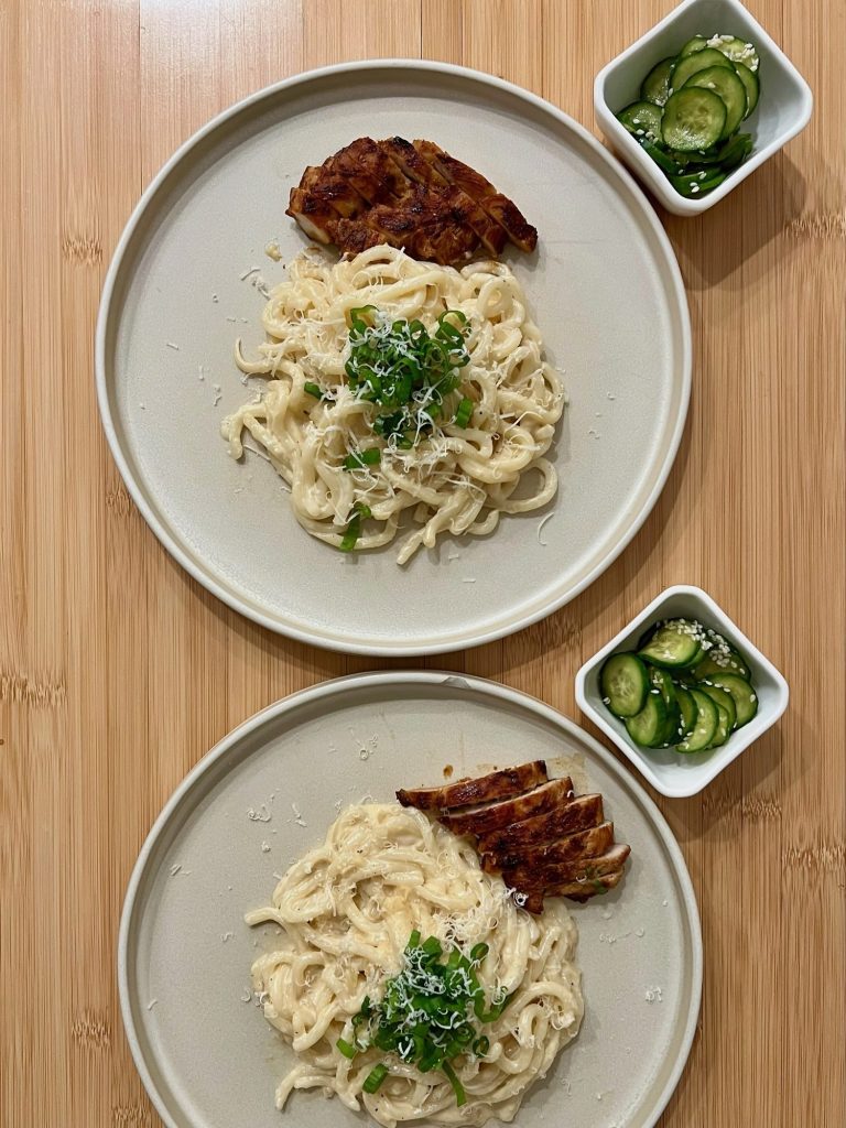 Two small dishes of Japanese cucumber salad served beside plates of creamy udon and grilled chicken