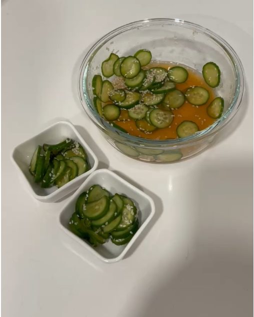 Sliced cucumbers marinating in rice vinegar and sesame seeds in a glass bowl, with two small dishes of cucumber salad