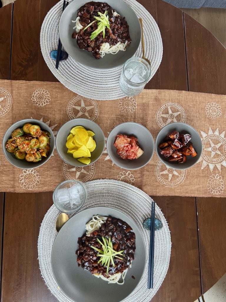 Overhead view of a table set with two plates of black bean noodles and small bowls of side dishes