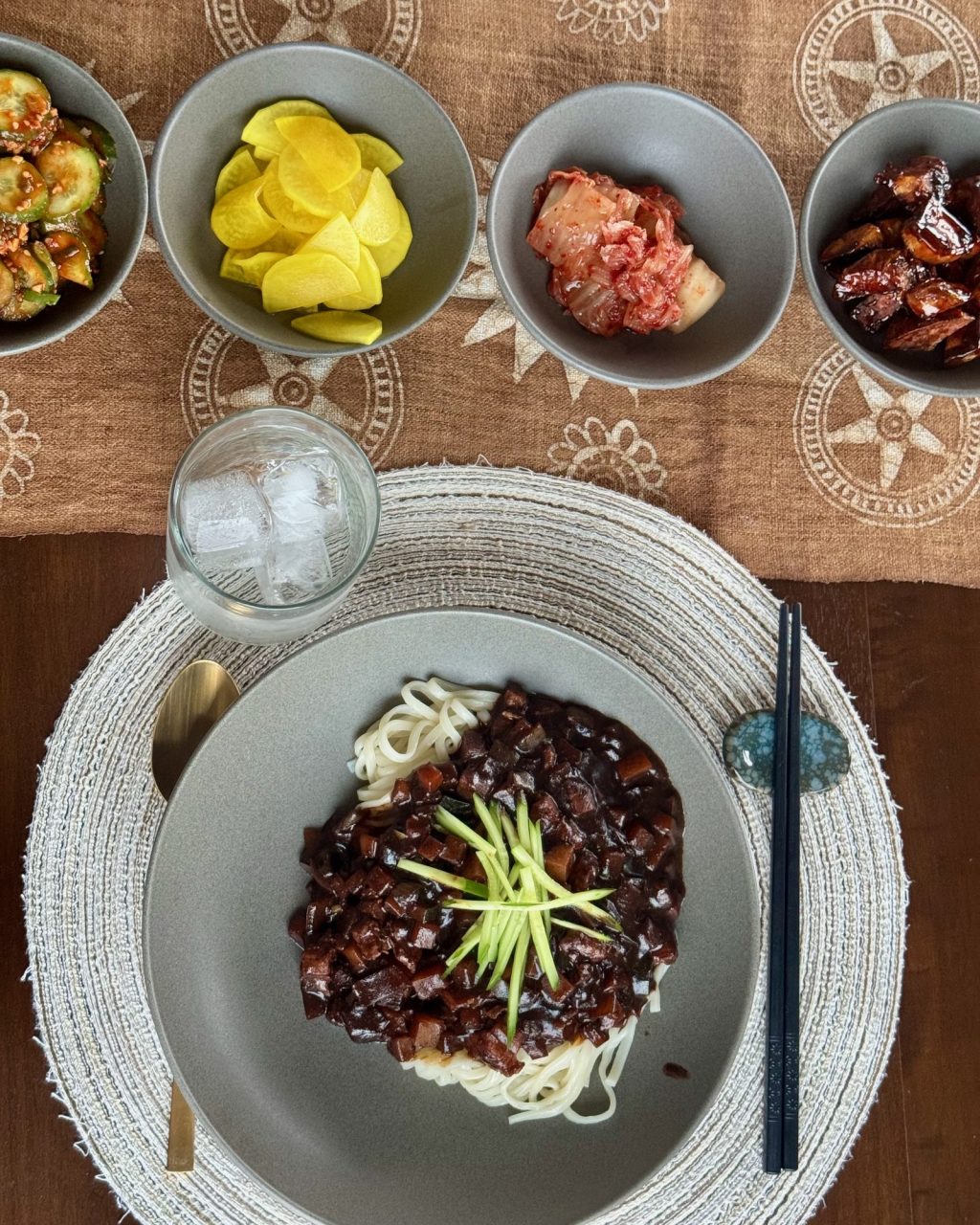 Plate of noodles topped with black bean sauce and cucumber matchsticks, with banchan bowls behind