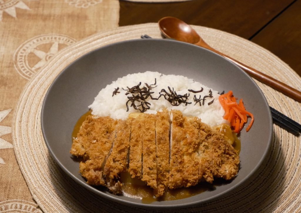 Sliced chicken katsu over Japanese curry with a mound of white rice and pickles on the side, with a wooden spoon on the placemat