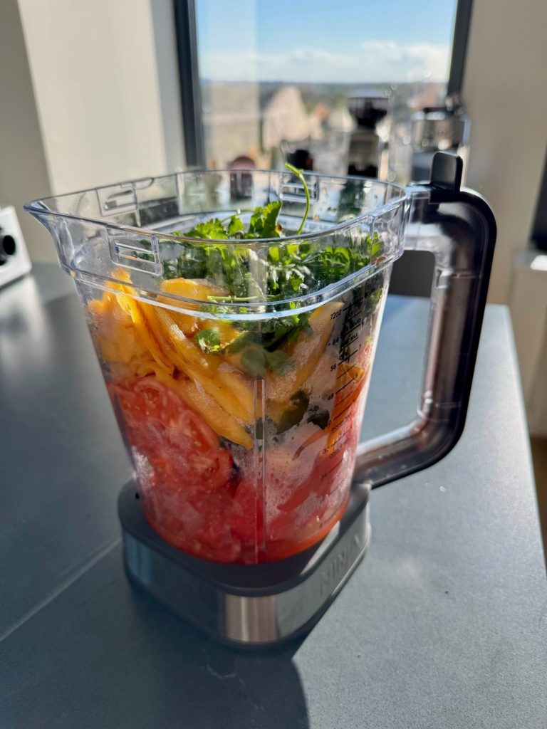 Blender filled with roasted tomatoes, peppers, browned mango and fresh cilantro, ready to blend