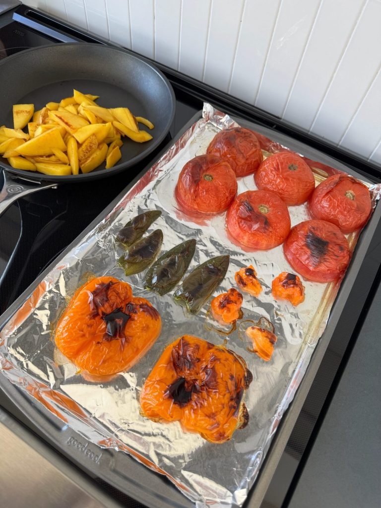 Charred tomatoes, bell peppers, jalapeños and habaneros on a foil-lined baking sheet next to a pan of browned mango