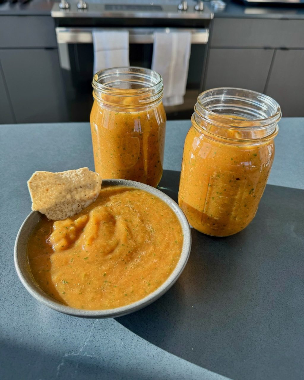 Bowl of orange roasted mango habanero salsa with a tortilla chip, next to two filled mason jars