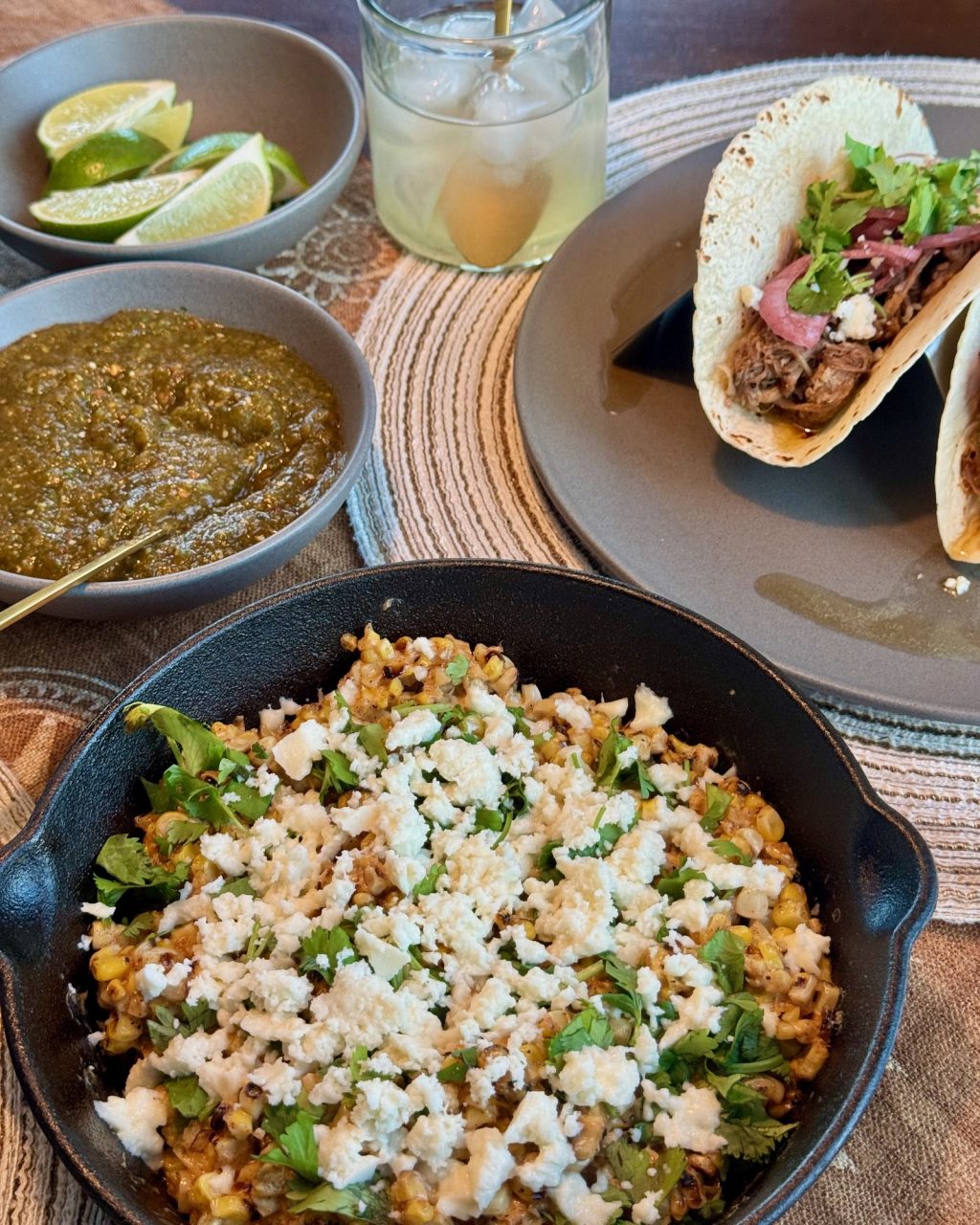 Cast iron skillet of charred corn kernels topped with crumbled queso fresco and cilantro, with tacos, salsa verde and lime wedges on the table