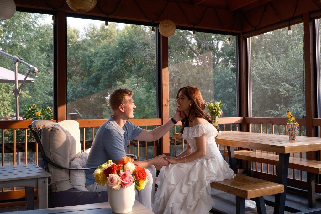Mia and Derrick sitting together on a screened porch at golden hour, with a vase of roses on the table