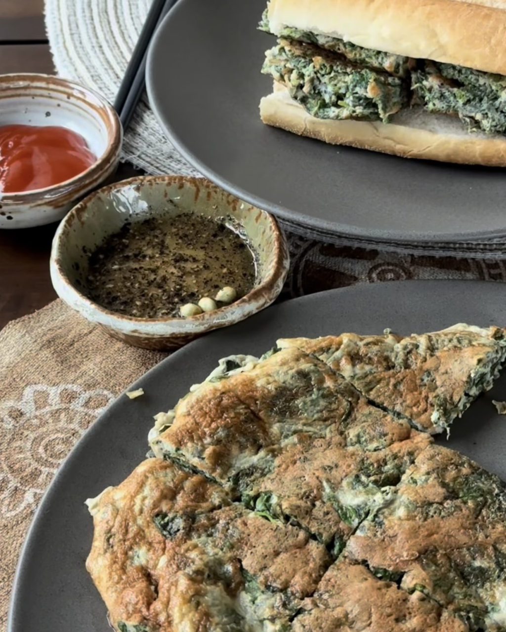 Mugwort omelette cut into wedges on a gray plate, with a salt, pepper and calamansi dip, chili sauce and a bánh mì stuffed with omelette behind