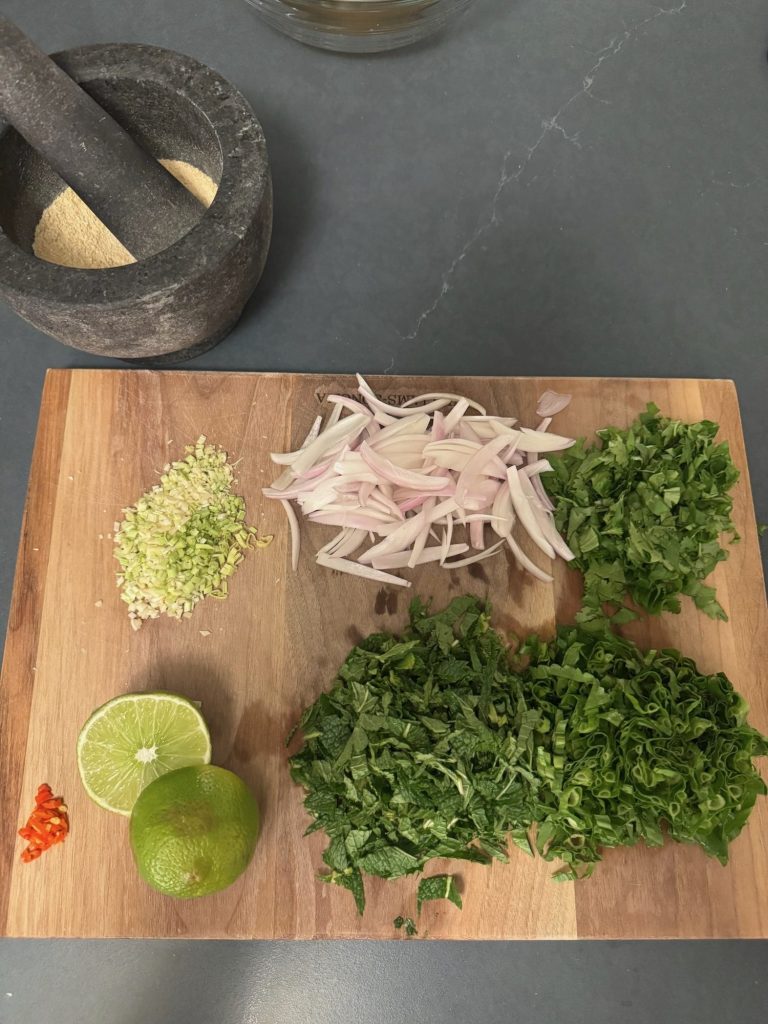 Cutting board with toasted rice powder, sliced shallots, cilantro, mint, limes and chili next to a mortar and pestle