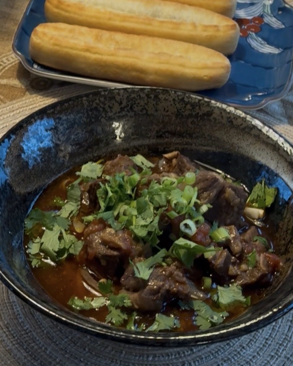 Dark bowl of red wine braised beef in a rich broth, topped with cilantro and green onion, with bread on a plate behind