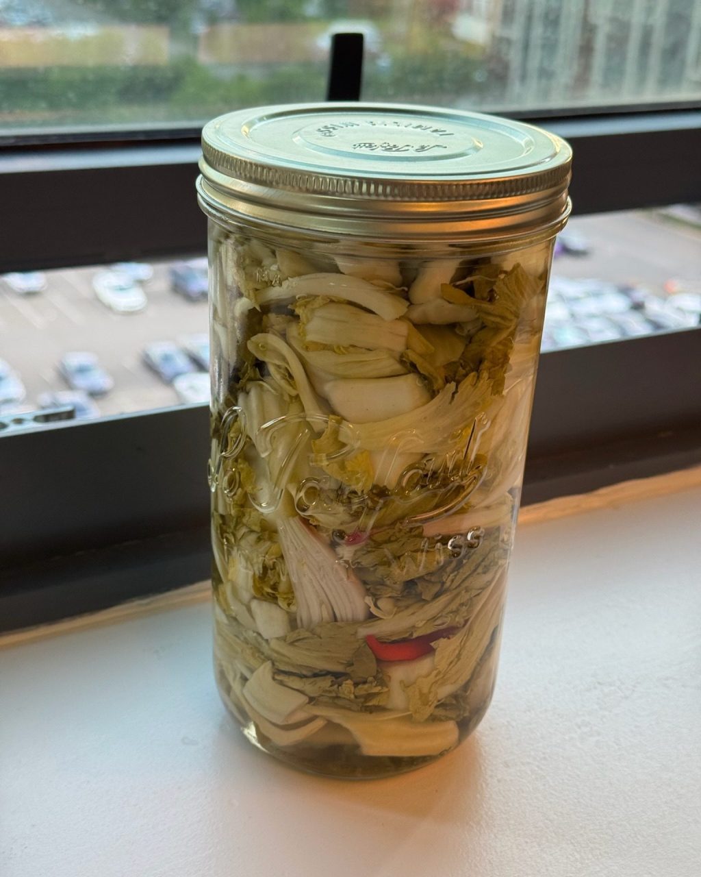 A tall jar of pickled mustard greens in brine on a windowsill