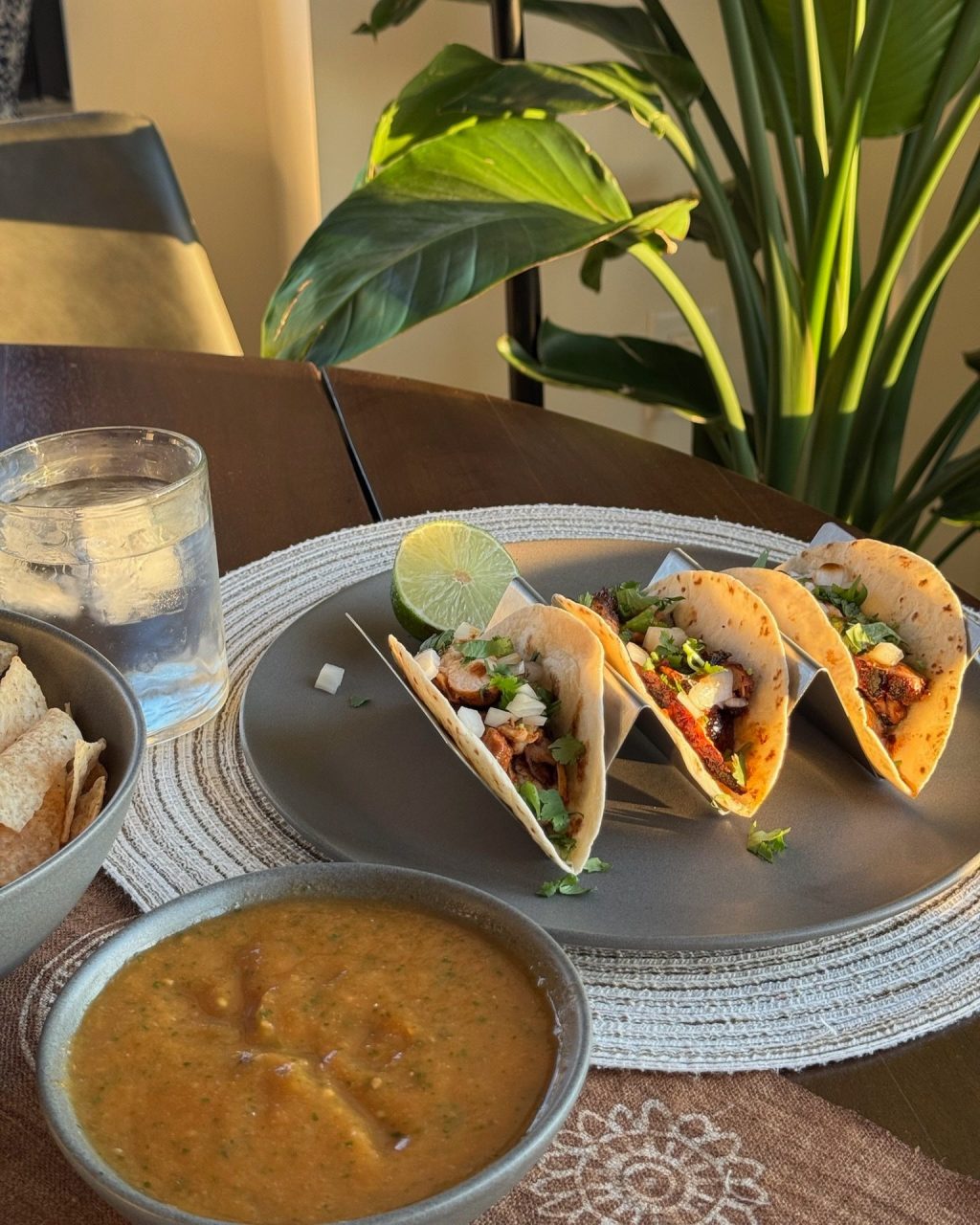 Three pollo asado tacos with onion and cilantro in a taco holder, with salsa, chips and a lime wedge on the table