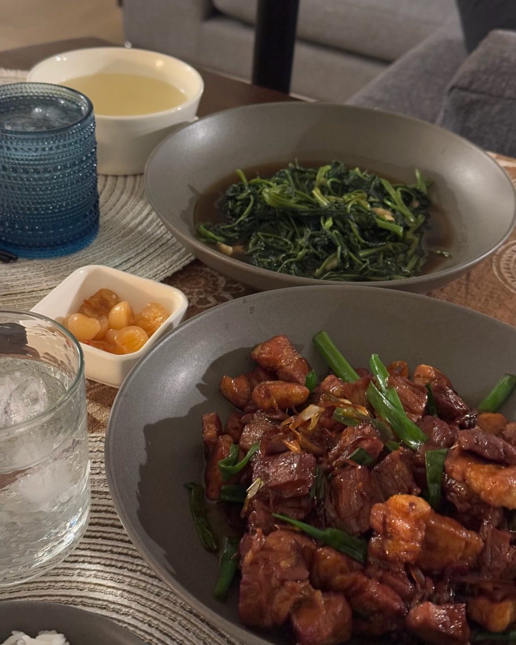 Stir-fried morning glory in a grey bowl beside a cup of broth, with a plate of caramelized pork and a dish of quail eggs on the table