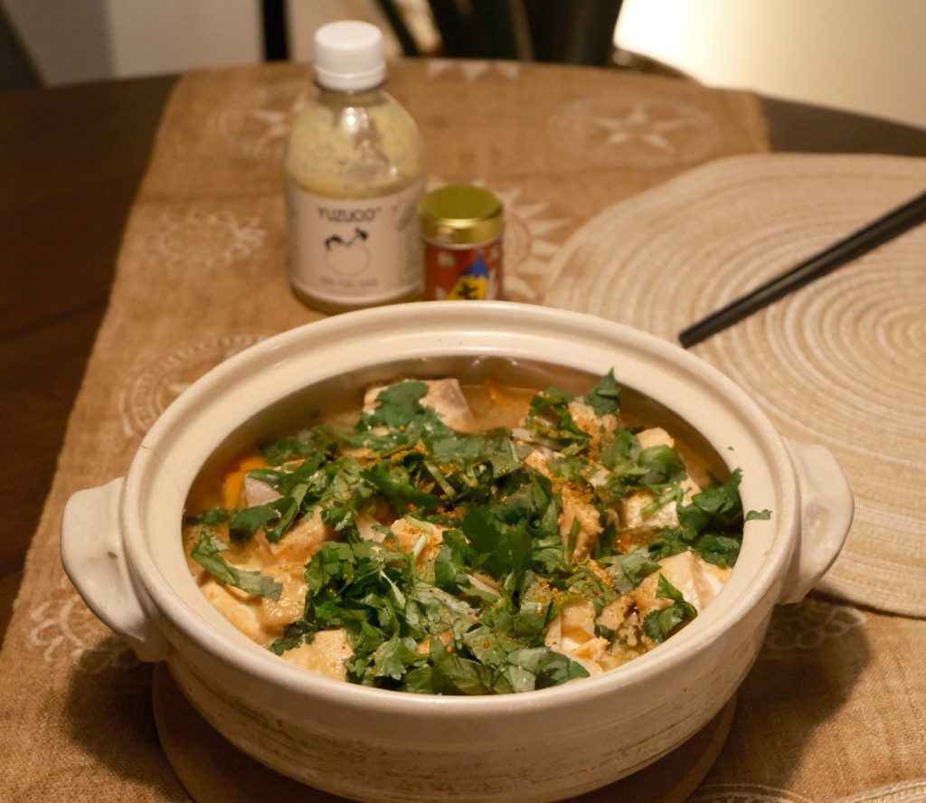 Donabe of salmon and daikon in a golden garlic butter miso broth, topped with fresh herbs and chili, with yuzu sauce and a bowl of rice on the table
