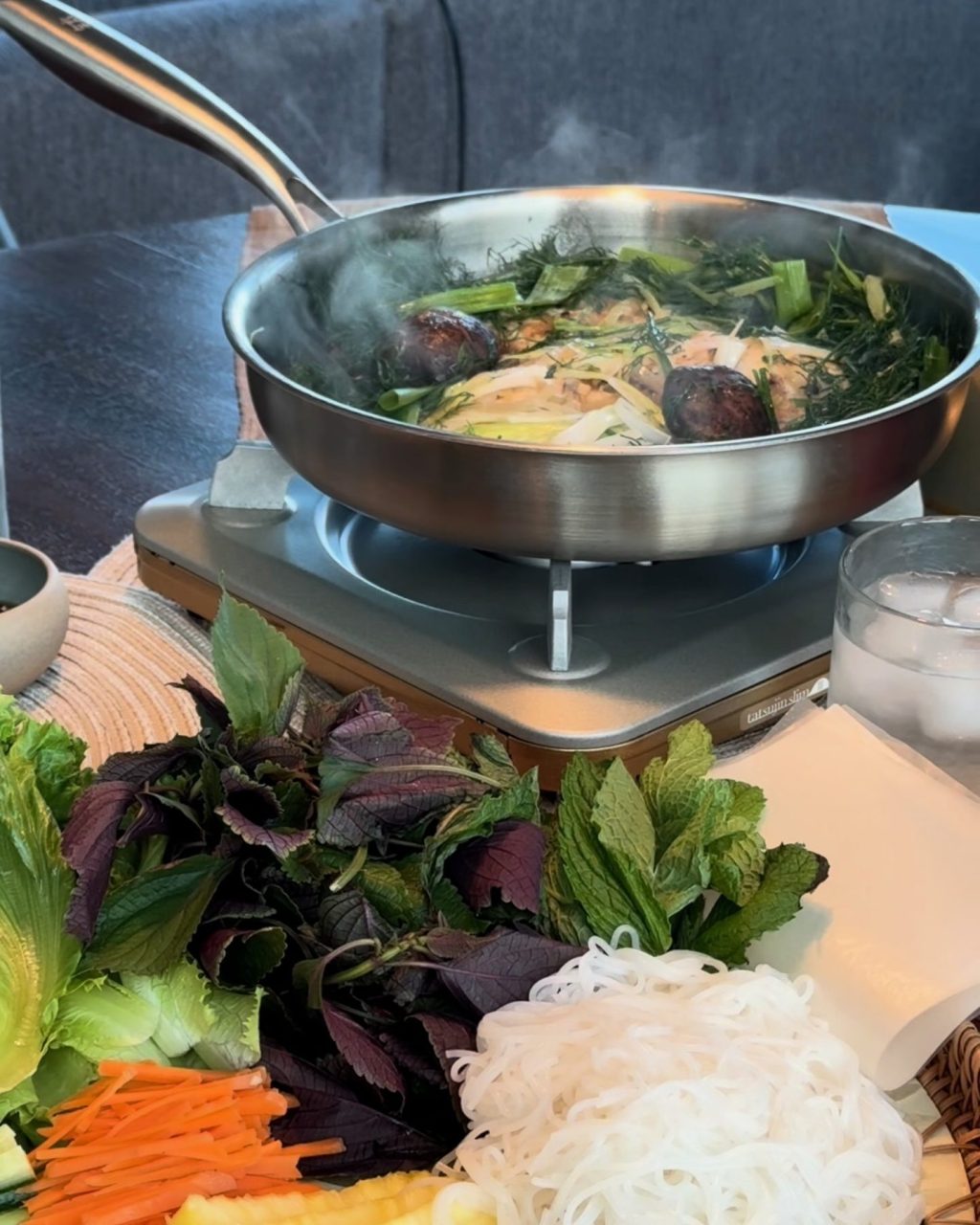Skillet of soy-steamed fish with shiitake, dill and green onion on a tabletop burner, with herbs, vermicelli, carrot and pineapple for rolling