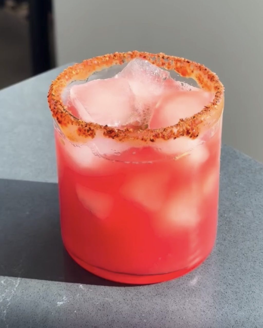 A glass of pink watermelon limeade over ice with a chamoy and Tajín rim, in bright sunlight