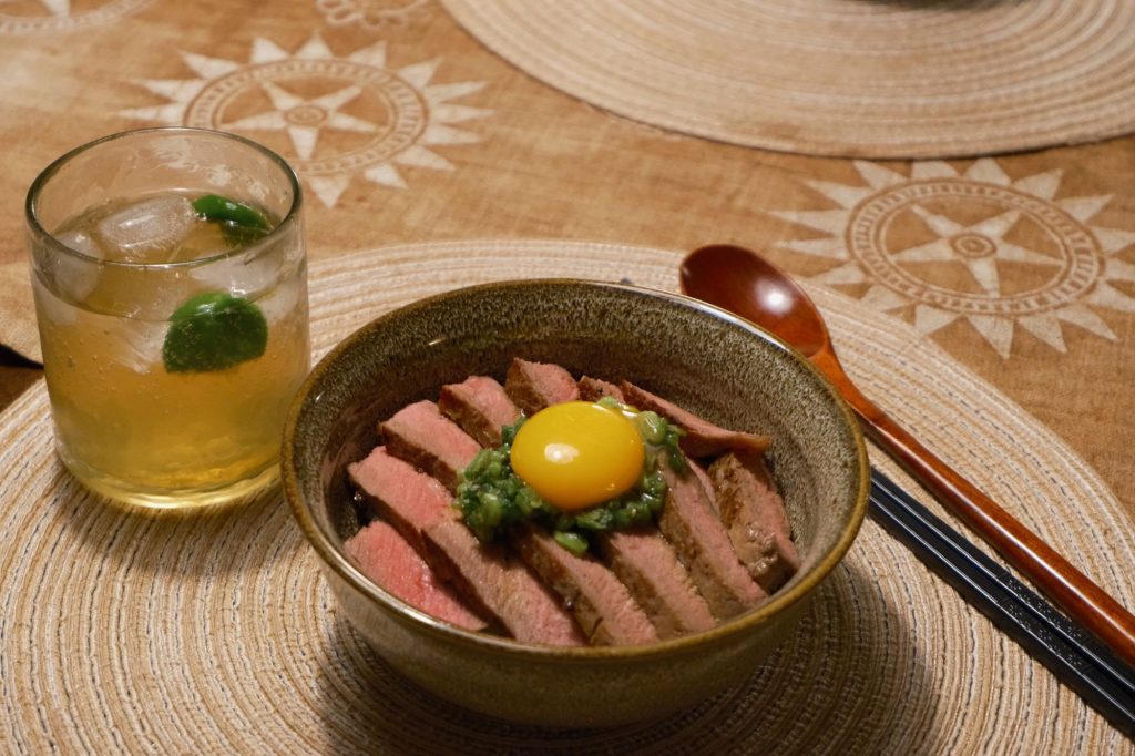 Bowl of rice topped with thinly sliced medium-rare steak, a yuzu kosho scallion sauce and a raw egg yolk, with an iced drink and a wooden spoon on the table
