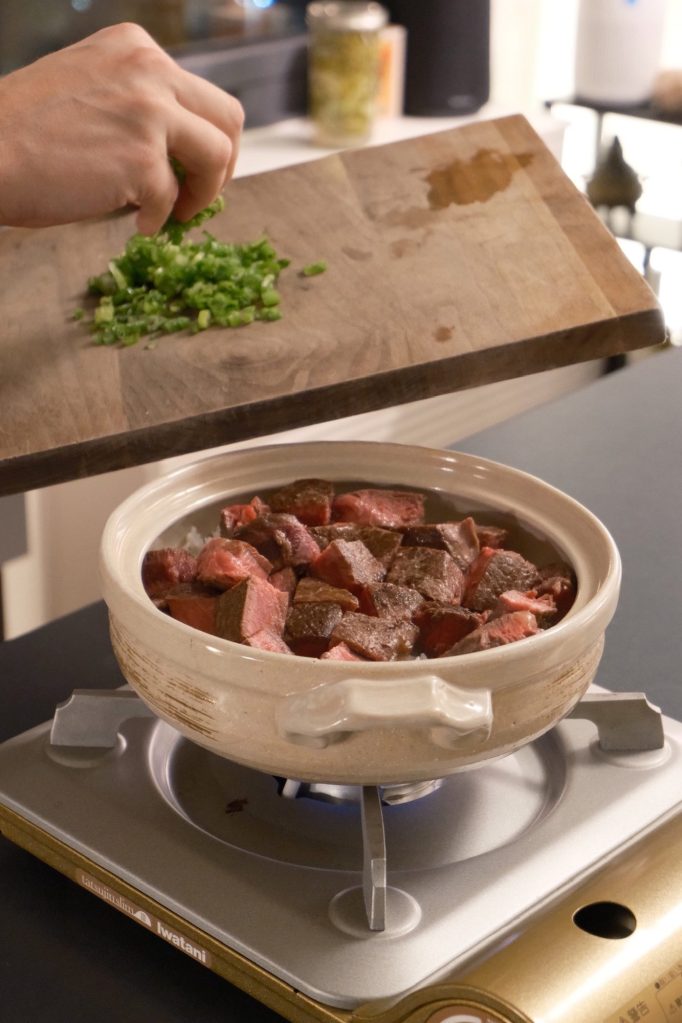 Sliced green onions being added from a cutting board to a donabe of cubed rare steak on a portable burner