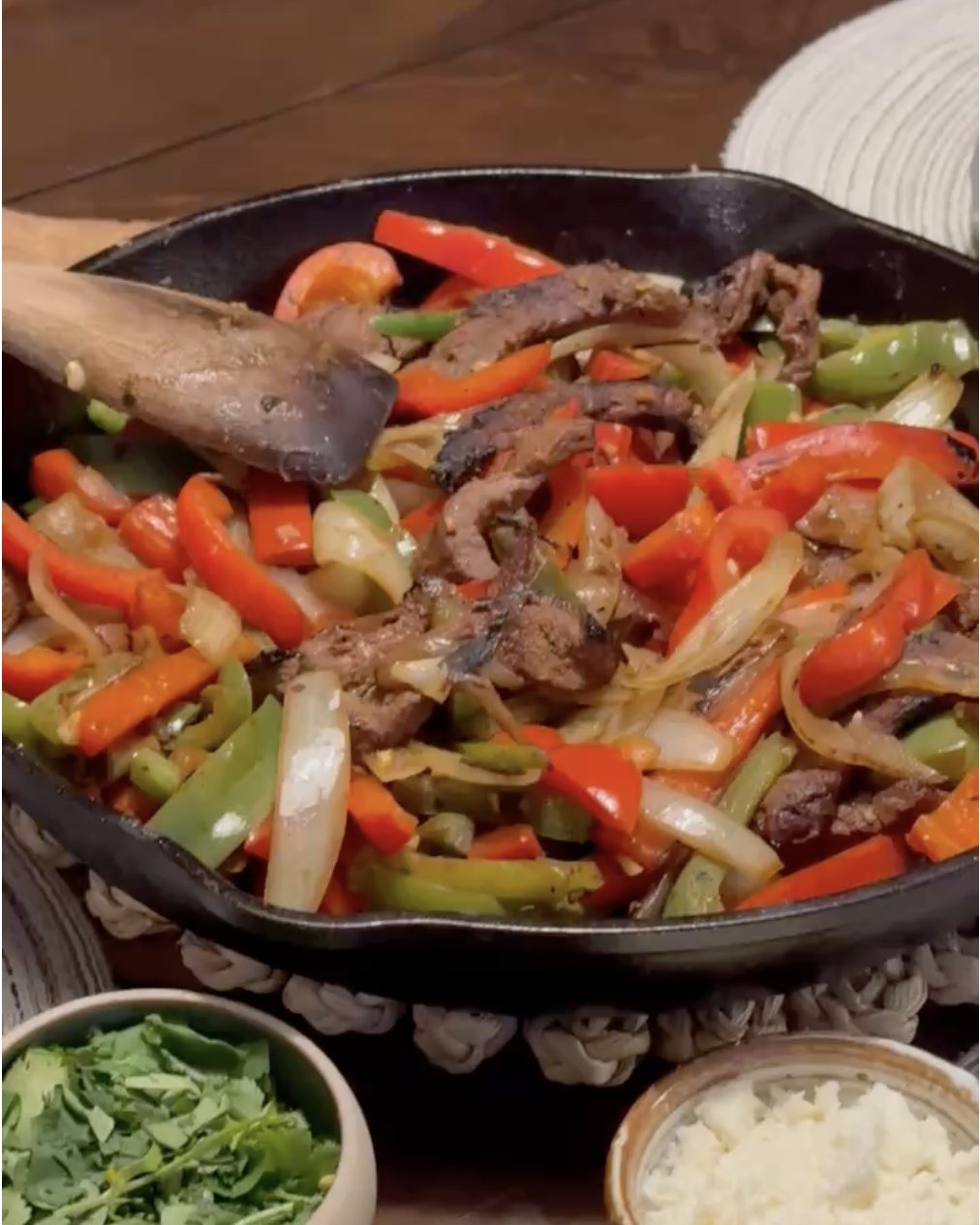Cast iron skillet of sliced skirt steak with red, orange and green bell peppers and onions, with cilantro, queso fresco and lime wedges