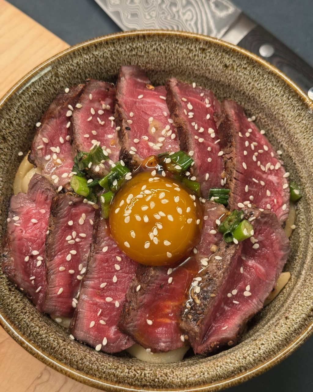 Bowl of udon topped with sliced rare steak, green onions, sesame seeds and a soy-cured egg yolk