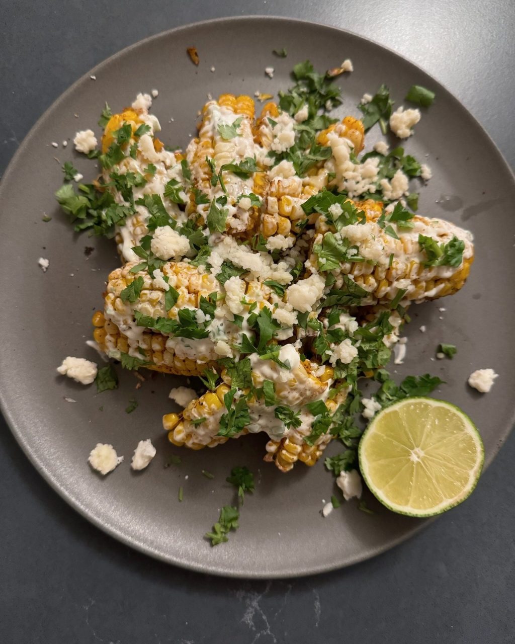 Corn ribs topped with crema, crumbled cotija and cilantro on a grey plate with a lime half