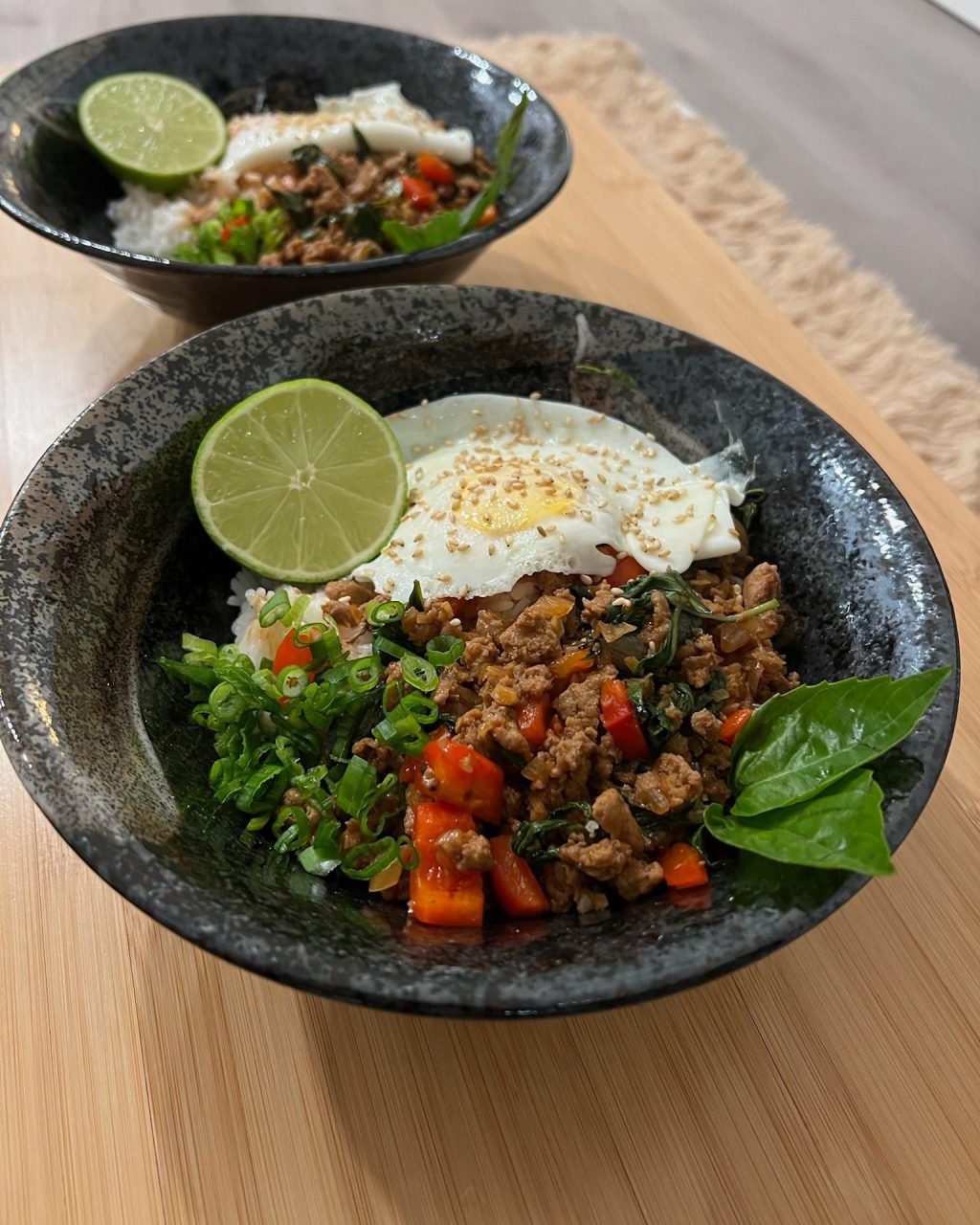 Thai basil chicken over jasmine rice in a dark speckled bowl, topped with a crispy fried egg, lime half, green onion and fresh basil leaves