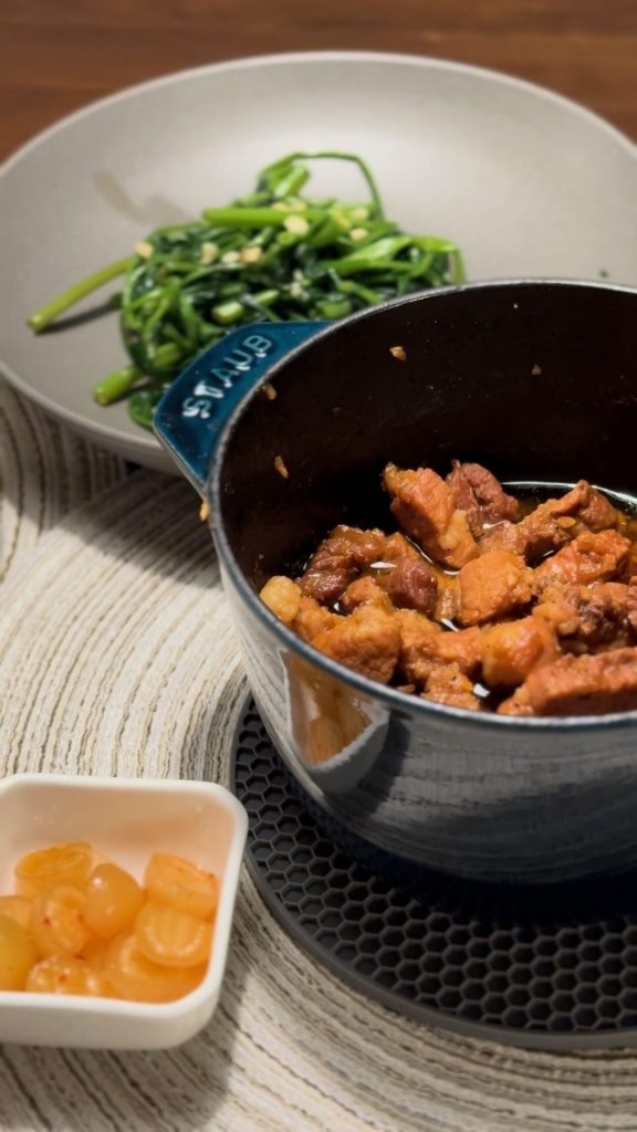 Caramelized pork belly braising in a dark blue Dutch oven, with a plate of stir-fried water spinach and a small dish of pickles on the table