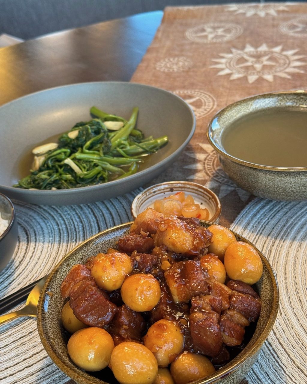 A bowl of thịt kho tàu: glossy caramelized pork belly braised with quail eggs, with stir-fried water spinach and a bowl of broth behind
