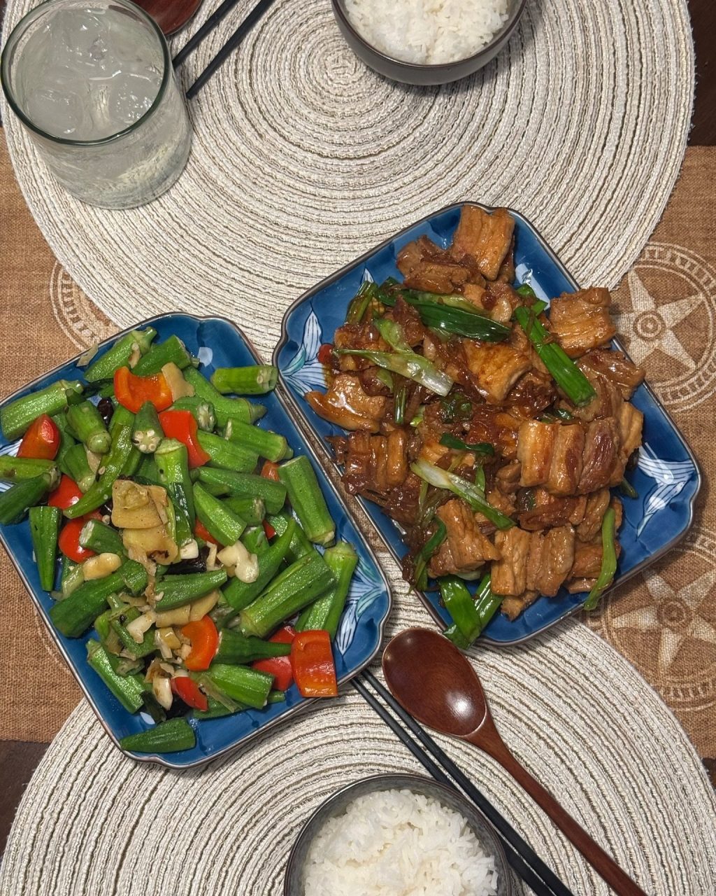 Overhead view of caramelized pork belly with green onion and garlicky stir-fried okra with red chili on blue plates, with bowls of white rice