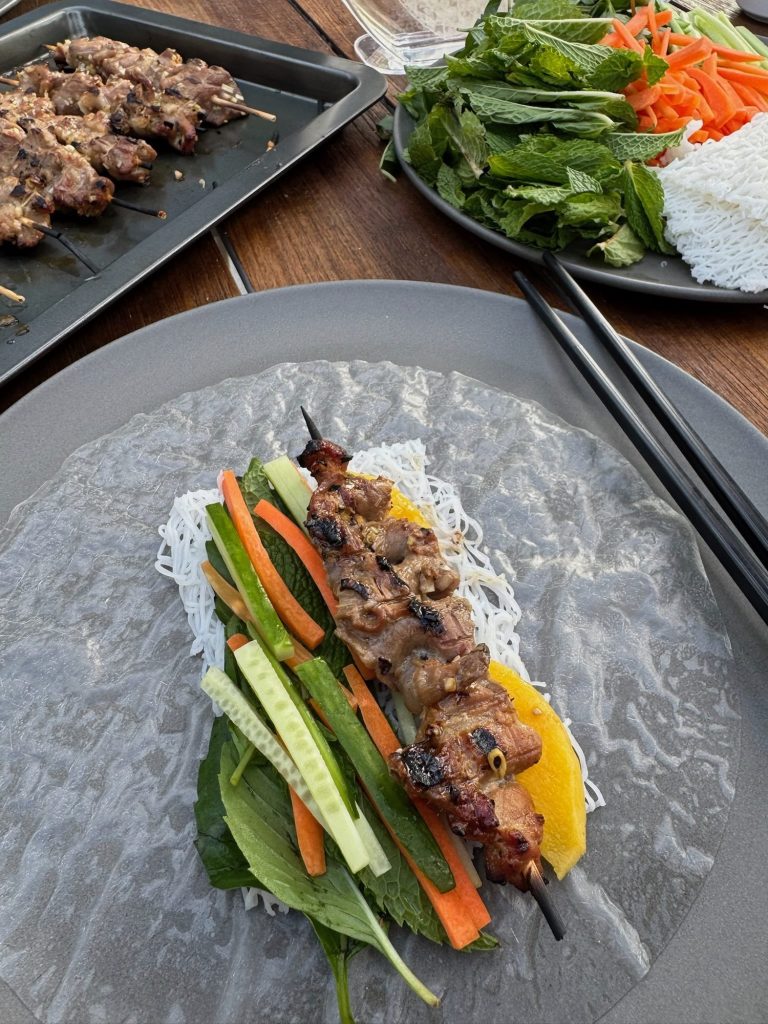 Rice paper laid flat with herbs, rice noodles, carrot, cucumber and a grilled pork skewer ready to roll