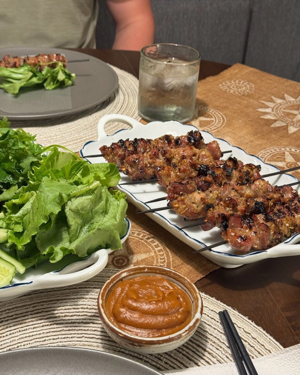 Grilled lemongrass pork skewers on a platter with lettuce, herbs, cucumber and a bowl of dipping sauce